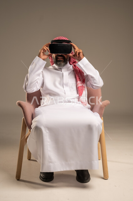 قضاء أوقات ممتعة ومسلية بالألعاب الافتراضية، الاستمتاع بمشاهدة أفلام الواقع الافتراضي، بورتريه لرجل عربي خليجي سعودي كبير بالسن يرتدي الثوب التقليدي و الشماغ يجلس على كرسي مريح ويرتدي نظارة الواقع الافتراضي ثلاثية الابعاد،  خلفية بيج