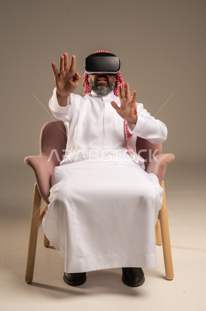 قضاء أوقات ممتعة ومسلية بالألعاب الافتراضية، الاستمتاع بمشاهدة أفلام الواقع الافتراضي، بورتريه لرجل عربي خليجي سعودي كبير بالسن يرتدي الثوب التقليدي و الشماغ يجلس على كرسي مريح ويرتدي نظارة الواقع الافتراضي ثلاثية الابعاد،  خلفية بيج