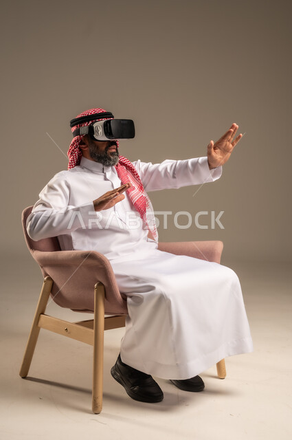 قضاء أوقات ممتعة ومسلية بالألعاب الافتراضية، الاستمتاع بمشاهدة أفلام الواقع الافتراضي، بورتريه لرجل عربي خليجي سعودي كبير بالسن يرتدي الثوب التقليدي و الشماغ يجلس على كرسي مريح ويرتدي نظارة الواقع الافتراضي ثلاثية الابعاد،  خلفية بيج