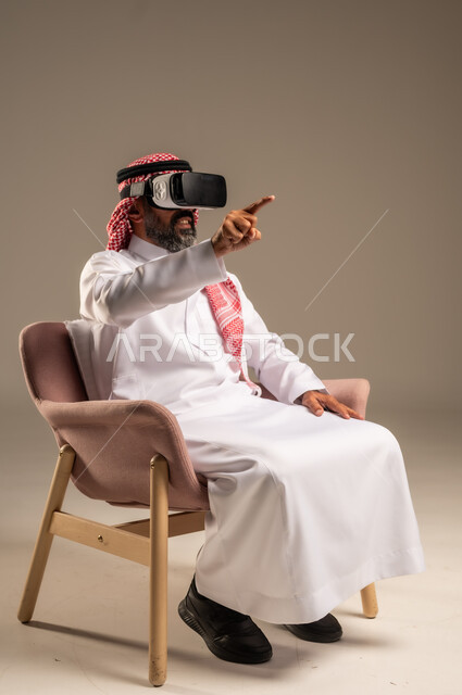 قضاء أوقات ممتعة ومسلية بالألعاب الافتراضية، الاستمتاع بمشاهدة أفلام الواقع الافتراضي، بورتريه لرجل عربي خليجي سعودي كبير بالسن يرتدي الثوب التقليدي و الشماغ يجلس على كرسي مريح ويرتدي نظارة الواقع الافتراضي ثلاثية الابعاد،  خلفية بيج