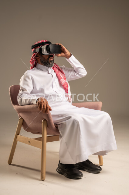 قضاء أوقات ممتعة ومسلية بالألعاب الافتراضية، الاستمتاع بمشاهدة أفلام الواقع الافتراضي، بورتريه لرجل عربي خليجي سعودي كبير بالسن يرتدي الثوب التقليدي و الشماغ يجلس على كرسي مريح ويرتدي نظارة الواقع الافتراضي ثلاثية الابعاد،  خلفية بيج