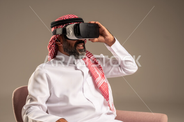 قضاء أوقات ممتعة ومسلية بالألعاب الافتراضية، الاستمتاع بمشاهدة أفلام الواقع الافتراضي، بورتريه لرجل عربي خليجي سعودي كبير بالسن يرتدي الثوب التقليدي و الشماغ يجلس على كرسي مريح ويرتدي نظارة الواقع الافتراضي ثلاثية الابعاد،  خلفية بيج