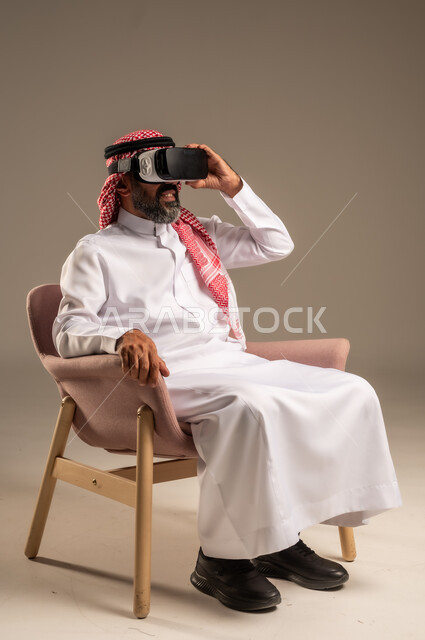 قضاء أوقات ممتعة ومسلية بالألعاب الافتراضية، الاستمتاع بمشاهدة أفلام الواقع الافتراضي، بورتريه لرجل عربي خليجي سعودي كبير بالسن يرتدي الثوب التقليدي و الشماغ يجلس على كرسي مريح ويرتدي نظارة الواقع الافتراضي ثلاثية الابعاد،  خلفية بيج