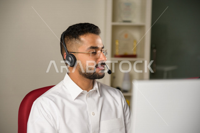 موظف عربي خليجي سعودي يعمل في خدمة العملاء ويرتدي سماعة الاتصال، استعلامات واستفسارات، الاجابه على اسئلة العملاء، اجراء اتصال فيديو من خلال الحاسوب، نظارة طبية، متابعة وتطوير العمل