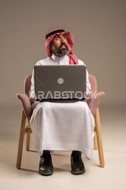 استخدام اللابتوب والعمل عن بعد، بورتريه مقرب لرجل عربي خليجي سعودي كبير بالسن يرتدي الشماغ والثوب التقليدي يجلس على الكرسي يحمل الحاسوب المحمول، تطور التكنولوجيا والأجهزة التقنية، خلفية بيج