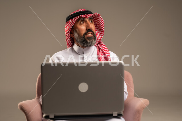 استخدام اللابتوب والعمل عن بعد، بورتريه مقرب لرجل عربي خليجي سعودي كبير بالسن يرتدي الشماغ والثوب التقليدي يجلس على الكرسي يحمل الحاسوب المحمول، تطور التكنولوجيا والأجهزة التقنية، خلفية بيج