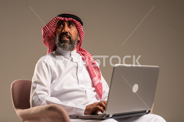 استخدام اللابتوب والعمل عن بعد، بورتريه مقرب لرجل عربي خليجي سعودي كبير بالسن يرتدي الشماغ والثوب التقليدي يجلس على الكرسي يحمل الحاسوب المحمول، تطور التكنولوجيا والأجهزة التقنية، خلفية بيج