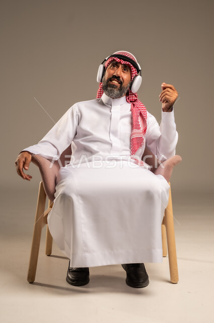 الإصغاء والاستمتاع باستخدام الهيدفون، قضاء أوقات ممتعة ومسلية، الاستماع إلى المواهب الغنائية الفنية، بورتريه لرجل عربي خليجي سعودي كبير بالسن يرتدي الثوب التقليدي والشماغ يضع سماعة الرأس ويستمتع بسماع اغاني منوعة، خلفية بيج