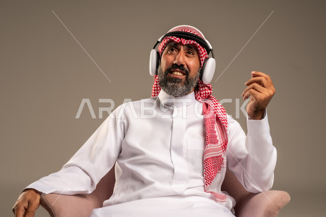 الإصغاء والاستمتاع باستخدام الهيدفون، قضاء أوقات ممتعة ومسلية، الاستماع إلى المواهب الغنائية الفنية، بورتريه لرجل عربي خليجي سعودي كبير بالسن يرتدي الثوب التقليدي والشماغ يضع سماعة الرأس ويستمتع بسماع اغاني منوعة، خلفية بيج