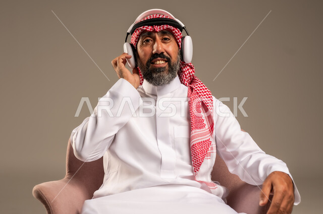 الإصغاء والاستمتاع باستخدام الهيدفون، قضاء أوقات ممتعة ومسلية، الاستماع إلى المواهب الغنائية الفنية، بورتريه لرجل عربي خليجي سعودي كبير بالسن يرتدي الثوب التقليدي والشماغ يضع سماعة الرأس ويستمتع بسماع اغاني منوعة، خلفية بيج