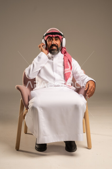الإصغاء والاستمتاع باستخدام الهيدفون، قضاء أوقات ممتعة ومسلية، الاستماع إلى المواهب الغنائية الفنية، بورتريه لرجل عربي خليجي سعودي كبير بالسن يرتدي الثوب التقليدي والشماغ يضع سماعة الرأس ويستمتع بسماع اغاني منوعة، خلفية بيج