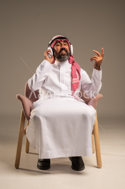 الإصغاء والاستمتاع باستخدام الهيدفون، قضاء أوقات ممتعة ومسلية، الاستماع إلى المواهب الغنائية الفنية، بورتريه لرجل عربي خليجي سعودي كبير بالسن يرتدي الثوب التقليدي والشماغ يضع سماعة الرأس ويستمتع بسماع اغاني منوعة، خلفية بيج