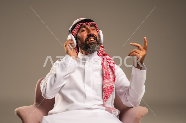 الإصغاء والاستمتاع باستخدام الهيدفون، قضاء أوقات ممتعة ومسلية، الاستماع إلى المواهب الغنائية الفنية، بورتريه لرجل عربي خليجي سعودي كبير بالسن يرتدي الثوب التقليدي والشماغ يضع سماعة الرأس ويستمتع بسماع اغاني منوعة، خلفية بيج