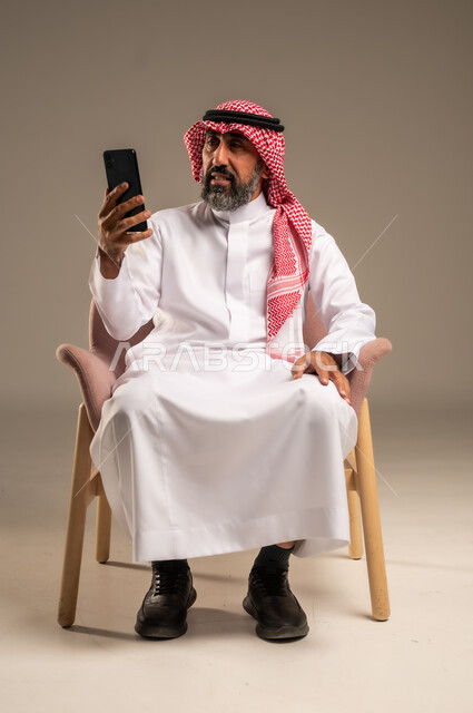 استخدام التطبيقات الحديثة و التسويق الألكتروني, بورتريه لرجل عربي سعودي خليجي كبير بالسن  يرتدي الثوب التقليدي والشماغ يستخدم الجوال, برامج التواصل الاجتماعي عبر الهاتف المحمول, خلفية بيج .