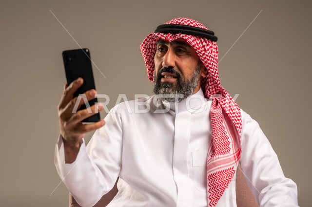 استخدام التطبيقات الحديثة و التسويق الألكتروني, بورتريه لرجل عربي سعودي خليجي كبير بالسن  يرتدي الثوب التقليدي والشماغ يستخدم الجوال, برامج التواصل الاجتماعي عبر الهاتف المحمول, خلفية بيج .