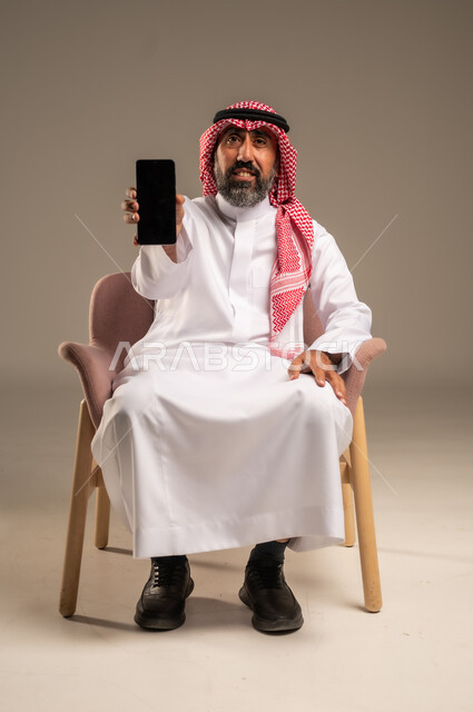 استخدام التطبيقات الحديثة و التسويق الألكتروني, بورتريه لرجل عربي سعودي خليجي كبير بالسن  يرتدي الثوب التقليدي والشماغ يمسك الجوال بشاشة سوداء فارغة, برامج التواصل الاجتماعي عبر الهاتف المحمول, خلفية بيج .