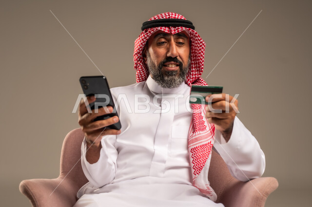 Online shopping and payment, using credit card in online shopping, portrait of an elderly Saudi Arabian Gulf man wearing a traditional keffiyeh and thobe sitting on a chair holding a bank card and browsing banking applications via mobile, beige background