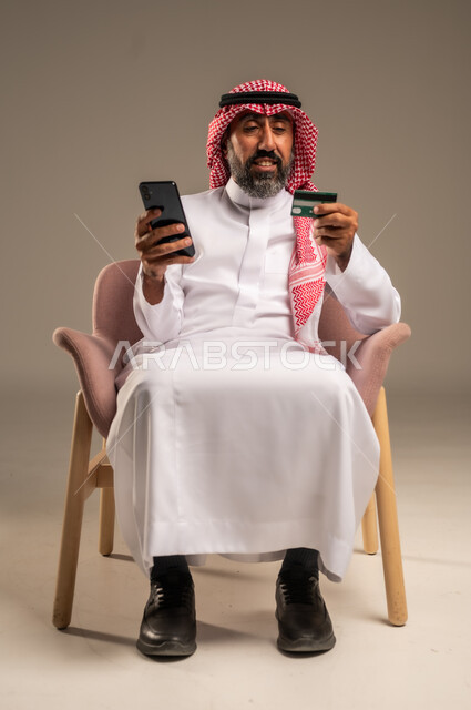 Online shopping and payment, using credit card in online shopping, portrait of an elderly Saudi Arabian Gulf man wearing a traditional keffiyeh and thobe sitting on a chair holding a bank card and browsing banking applications via mobile, beige background