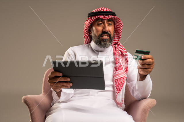 الشراء والدفع عن بعد عبر الاونلاين, استخدام بطاقة الإئتمان في التسوق الالكتروني، بورتريه مقرب لرجل عربي سعودي خليجي كبير بالسن يرتدي الشماغ والثوب التقليدي يجلس على كرسي يمسك البطاقة البنكية ويقوم بتصفح تطبيقات المصارف والبنوك عبر التابلت، خلفية بيج