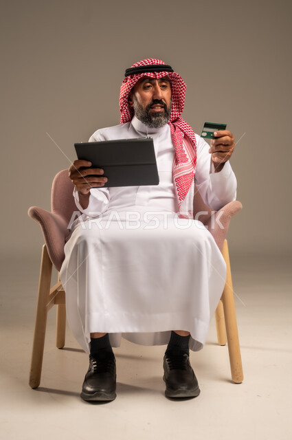 Online shopping and payment, using credit card in online shopping, close-up portrait of elderly Saudi Arabian Gulf man wearing traditional keffiyeh and thobe sitting on chair holding bank card and browsing banking applications on tablet, beige background