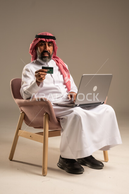 Online shopping and payment, using credit card in online shopping, portrait of an elderly Saudi Arabian Gulf man wearing a traditional keffiyeh and thobe sitting on a chair holding a bank card and browsing banking applications on a laptop, beige background