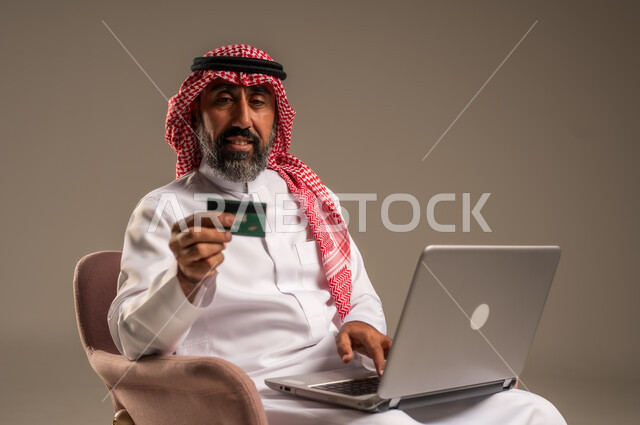 الشراء والدفع عن بعد عبر الاونلاين, استخدام بطاقة الإئتمان في التسوق الالكتروني، بورتريه  لرجل عربي سعودي خليجي كبير بالسن يرتدي الشماغ والثوب التقليدي يجلس على كرسي يمسك البطاقة البنكية ويقوم بتصفح تطبيقات المصارف والبنوك عبر اللابتوب، خلفية بيج