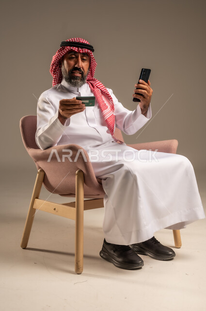 الشراء والدفع عن بعد عبر الاونلاين, استخدام بطاقة الإئتمان في التسوق الالكتروني، بورتريه  لرجل عربي سعودي خليجي كبير بالسن يرتدي الشماغ والثوب التقليدي يجلس على كرسي يمسك البطاقة البنكية ويقوم بتصفح تطبيقات المصارف والبنوك عبر الجوال، خلفية بيج