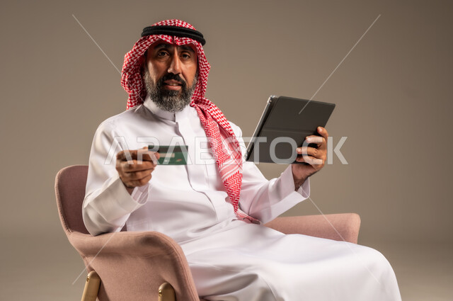 الشراء والدفع عن بعد عبر الاونلاين, استخدام بطاقة الإئتمان في التسوق الالكتروني، بورتريه مقرب لرجل عربي سعودي خليجي كبير بالسن يرتدي الشماغ والثوب التقليدي يجلس على كرسي يمسك البطاقة البنكية ويقوم بتصفح تطبيقات المصارف والبنوك عبر التابلت، خلفية بيج
