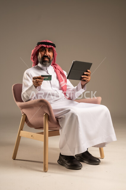 الشراء والدفع عن بعد عبر الاونلاين, استخدام بطاقة الإئتمان في التسوق الالكتروني، بورتريه مقرب لرجل عربي سعودي خليجي كبير بالسن يرتدي الشماغ والثوب التقليدي يجلس على كرسي يمسك البطاقة البنكية ويقوم بتصفح تطبيقات المصارف والبنوك عبر التابلت، خلفية بيج