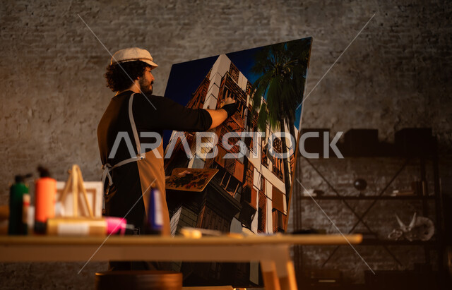 أنشطة ترفيهية ممتعة, ورشة عمل فنية , الشغف في ممارسة الهوايات المفضلة و الفنون اليدوية , صورة من الخلف لرسام عربي خليجي سعودي يرتدي الزي الكاجوال و مريلة الرسم يجلس على الكرسي أمام اللوحة، معرض الرسومات و اللوحات التشكيلية في المملكة العربية السعودية 