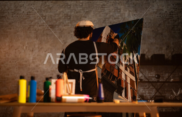 أنشطة ترفيهية ممتعة, ورشة عمل فنية , الشغف في ممارسة الهوايات المفضلة و الفنون اليدوية , صورة من الخلف لرسام عربي خليجي سعودي يرتدي الزي الكاجوال و مريلة الرسم يجلس على الكرسي أمام اللوحة، معرض الرسومات و اللوحات التشكيلية في المملكة العربية السعودية 