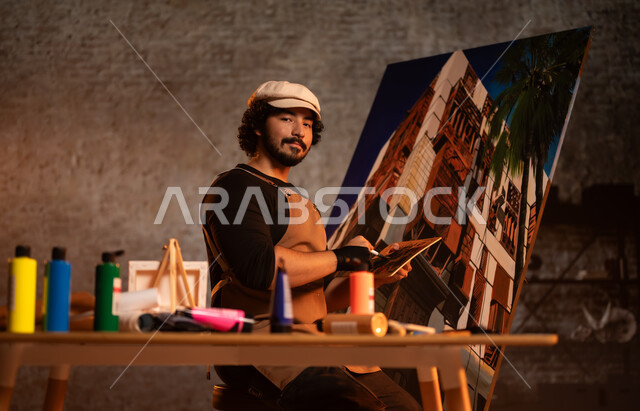 الشغف في ممارسة الهوايات المفضلة و الفنون اليدوية , أنشطة ترفيهية ممتعة, ورشة عمل فنية , صورة لرسام عربي خليجي سعودي يرتدي الزي الكاجوال و مريلة الرسم يجلس على الكرسي أمام اللوحة، معرض الرسومات و اللوحات التشكيلية في المملكة العربية السعودية 