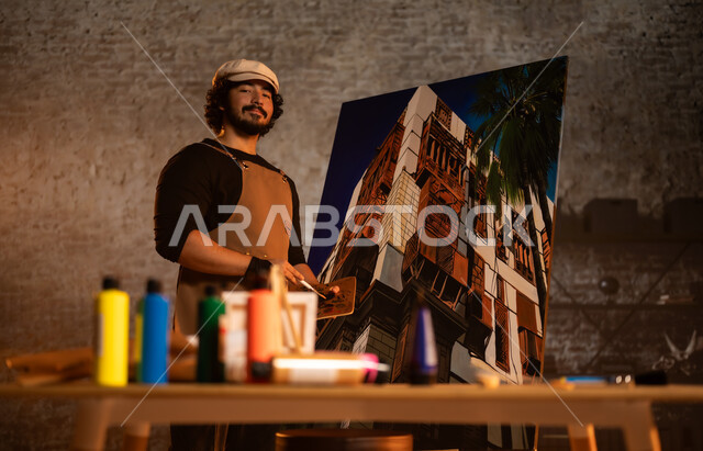 الشغف في ممارسة الهوايات المفضلة و الفنون اليدوية , أنشطة ترفيهية ممتعة, ورشة عمل فنية , صورة لرسام عربي خليجي سعودي يرتدي الزي الكاجوال و مريلة الرسم يجلس على الكرسي أمام اللوحة، معرض الرسومات و اللوحات التشكيلية في المملكة العربية السعودية 