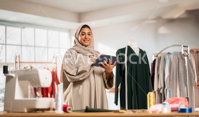 استخدام الاجهزة الالكترونية التقنية الحديثة، ورشة عمل تصميم أزياء, ممارسة مهنة الخياطة , مصممة أزياء عربية خليجية ترتدي العباءة الملونة والحجاب تمسك بيدها الجهاز اللوحي , حرف و مهن يدوية في المملكة، أعمال تقليدية شعبية, اتقان صنع الملابس السعودية