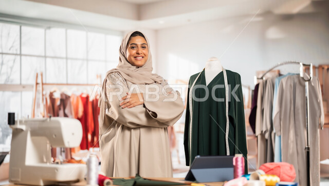 حرف و مهن يدوية في المملكة،  ورشة عمل تصميم أزياء, ممارسة مهنة الخياطة , مصممة أزياء عربية خليجية ترتدي العباءة الملونة والحجاب تقف بأيدي متقاطعة بايماءات السعادة والسرور, أعمال تقليدية شعبية, اتقان صنع الملابس السعودية
