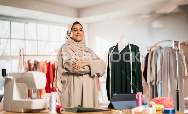 حرف و مهن يدوية في المملكة،  ورشة عمل تصميم أزياء, ممارسة مهنة الخياطة , مصممة أزياء عربية خليجية ترتدي العباءة الملونة والحجاب تقف بأيدي متقاطعة بايماءات السعادة والسرور, أعمال تقليدية شعبية, اتقان صنع الملابس السعودية
