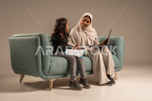 متابعة منصات التعليم الالكتروني المتاحة عبر الانترنت، دمج التعليم مع التقنية و التكنولوجيا، حل الواجبات المنزليه، معلمة خصوصي لمتابعة الدروس في المنزل، بورتريه لأم عربية خليجة سعودية محجبة تقوم بتعليم وتدريس ابنتها باستخدام الحاسب المحمول، خلفية رمادية