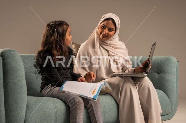 Follow up on e-learning platforms available online, integrating education with technology, solving homework, a private teacher to follow up on lessons at home, a portrait of a veiled Saudi Arabian Gulf mother teaching and educating her daughter using a laptop, gray background