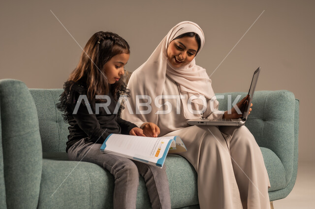 متابعة منصات التعليم الالكتروني المتاحة عبر الانترنت، دمج التعليم مع التقنية و التكنولوجيا، حل الواجبات المنزليه، معلمة خصوصي لمتابعة الدروس في المنزل، بورتريه لأم عربية خليجة سعودية محجبة تقوم بتعليم وتدريس ابنتها باستخدام الحاسب المحمول، خلفية رمادية