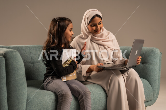 متابعة منصات التعليم الالكتروني المتاحة عبر الانترنت، دمج التعليم مع التقنية و التكنولوجيا، حل الواجبات المنزليه، معلمة خصوصي لمتابعة الدروس في المنزل، بورتريه لأم عربية خليجة سعودية محجبة تقوم بتعليم وتدريس ابنتها باستخدام الحاسب المحمول، خلفية رمادية