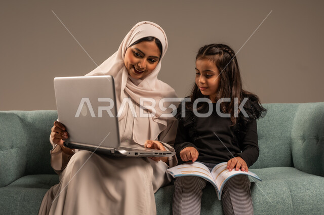 Integrating education with technology and technology, solving homework, a private teacher to follow up on lessons at home, following up on e-learning platforms available on the Internet, a portrait of a veiled Saudi Arabian Gulf mother teaching and educating her daughter using a laptop, gray background