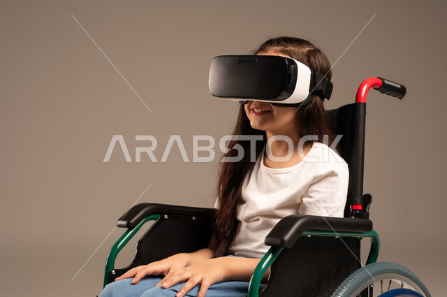  تحسين جودة حياة ذوي الاعاقة الحركية، استخدام كراسي المعاقين للمساعدة على الحركة والتنقل، بورتريه لفتاة عربية  خليجية سعودية ترتدي الزي الكاجوال تقوم بمشاهدة أفلام عبر نظارات الواقع الافتراضي ثلاثية الأبعاد، محاكاة العالم الخيالي والتفاعل مع الفيلم، خلفية رمادية