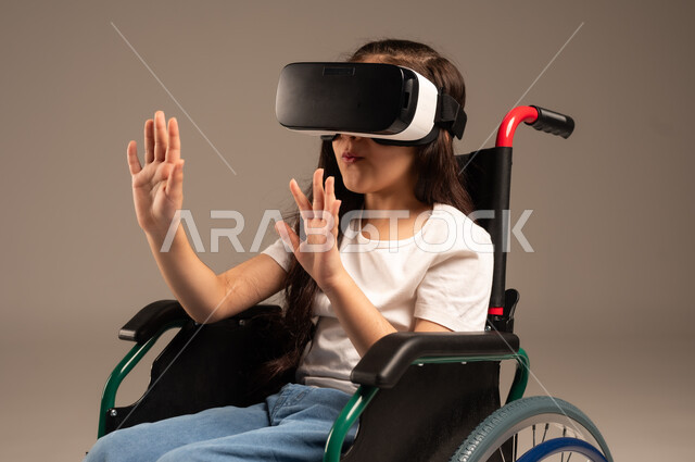  تحسين جودة حياة ذوي الاعاقة الحركية، استخدام كراسي المعاقين للمساعدة على الحركة والتنقل، بورتريه لفتاة عربية  خليجية سعودية ترتدي الزي الكاجوال تقوم بمشاهدة أفلام عبر نظارات الواقع الافتراضي ثلاثية الأبعاد، محاكاة العالم الخيالي والتفاعل مع الفيلم، خلفية رمادية