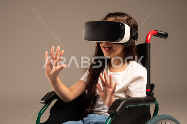  تحسين جودة حياة ذوي الاعاقة الحركية، استخدام كراسي المعاقين للمساعدة على الحركة والتنقل، بورتريه لفتاة عربية  خليجية سعودية ترتدي الزي الكاجوال تقوم بمشاهدة أفلام عبر نظارات الواقع الافتراضي ثلاثية الأبعاد، محاكاة العالم الخيالي والتفاعل مع الفيلم، خلفية رمادية