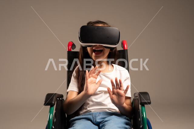 استخدام كراسي المعاقين للمساعدة على الحركة والتنقل،  تحسين جودة حياة ذوي الاعاقة الحركية، بورتريه لفتاة عربية  خليجية سعودية ترتدي الزي الكاجوال تقوم بمشاهدة أفلام عبر نظارات الواقع الافتراضي ثلاثية الأبعاد، محاكاة العالم الخيالي والتفاعل مع الفيلم، خلفية رمادية