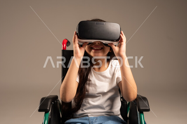استخدام كراسي المعاقين للمساعدة على الحركة والتنقل،  تحسين جودة حياة ذوي الاعاقة الحركية، بورتريه لفتاة عربية  خليجية سعودية ترتدي الزي الكاجوال تقوم بمشاهدة أفلام عبر نظارات الواقع الافتراضي ثلاثية الأبعاد، محاكاة العالم الخيالي والتفاعل مع الفيلم، خلفية رمادية