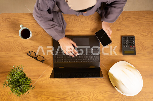 Supervising and auditing engineering projects and plans, using calculators to perform calculations, integrating technology and modern devices into practical life, working in accounting management, vertical upper portrait of an Arab Gulf Omani woman using a phone and a laptop
