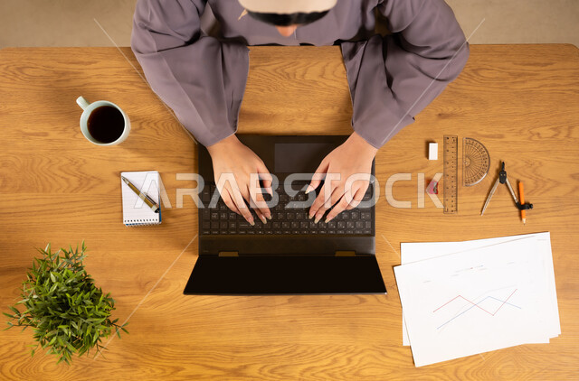 Online arithmetic training, learning arithmetic and mathematics through websites, using modern and advanced technologies, integrating technology into scientific life, vertical upper portrait of an Arab Gulf Omani woman using a laptop and next to her engineering tools