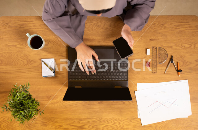 Using modern and advanced technologies, learning arithmetic and mathematics through websites, online arithmetic training, integrating technology into scientific life, vertical upper portrait of an Arab Gulf Omani woman using a phone and a laptop, displaying a blank black screen