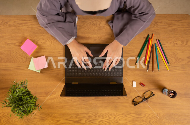 انجاز مهام العمل عن بعد، تدوين الأفكار وتسجيل المعلومات، تثبيت أوراق الملاحظات التذكارية الملونة، استخدام الأجهزة التقنية الحديثة  في الأعمال المنزلية، بورتريه علوي عامودي لإمرأة عربية خليجية عمانية تستخدم الحاسوب المحمول وبجانبها الألوان والنظارات الطبية